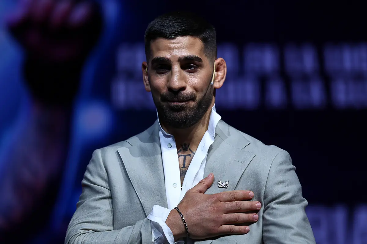 Hombre con barba y cabello corto vestido con traje gris claro y camisa blanca con la mano derecha sobre el pecho en un escenario oscuro