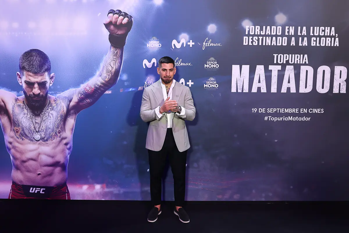 Hombre posando frente a un cartel promocional de una película titulada Topuria Matador con un luchador de artes marciales mixtas en el fondo y varios logotipos de patrocinadores Hombre posando frente a un cartel promocional de una película titulada Topuria Matador con un luchador de artes marciales mixtas en el fondo y varios logotipos de patrocinadores