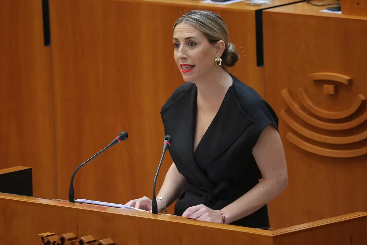 Mujer con vestido negro hablando en un atril de madera con dos micrófonos en un parlamento o sala de plenos