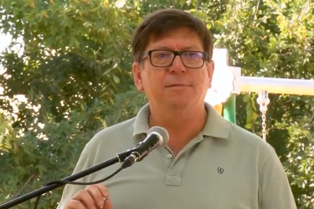 Hombre con gafas y polo verde hablando frente a un micrófono en un entorno al aire libre con árboles de fondo Hombre con gafas y polo verde hablando frente a un micrófono en un entorno al aire libre con árboles de fondo