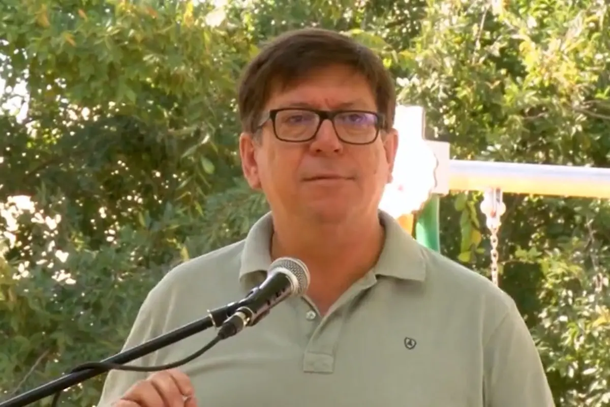 Hombre de cabello corto y gafas hablando frente a un micrófono al aire libre con árboles de fondo
