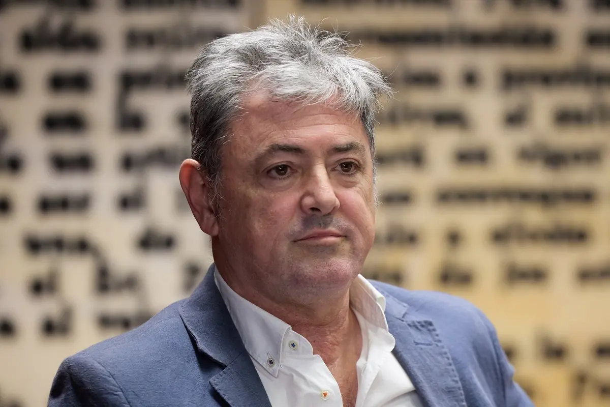 Hombre de cabello canoso y expresión seria viste saco azul claro y camisa blanca con fondo desenfocado de texto en letras negras