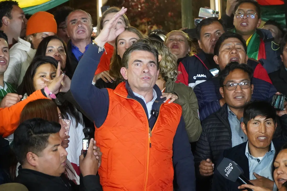 Hombre con chaleco naranja levantando la mano y hablando ante una multitud de personas que lo rodean en un evento público
