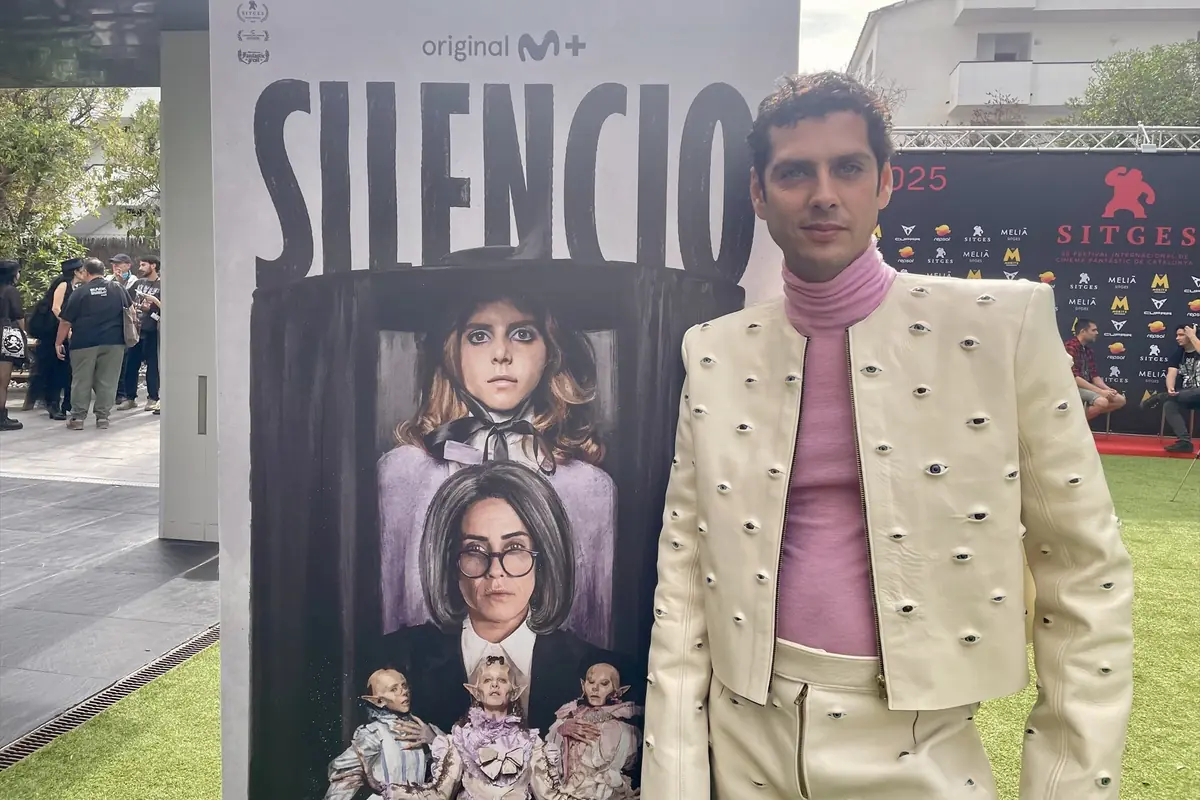 Hombre con traje blanco decorado con ojos y suéter rosa posando junto a un cartel de la serie Silencio en un evento al aire libre Hombre con traje blanco decorado con ojos y suéter rosa posando junto a un cartel de la serie Silencio en un evento al aire libre