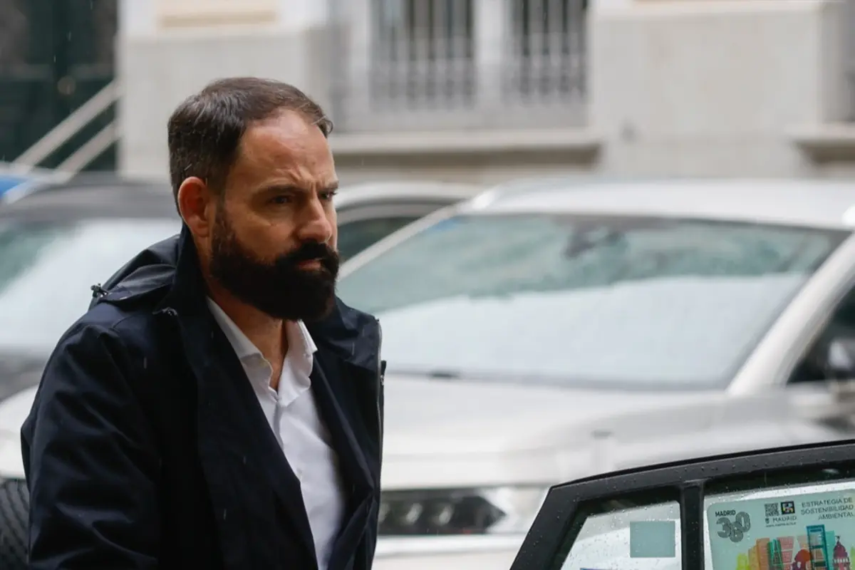 Hombre con barba y cabello corto usando abrigo oscuro junto a un coche en un día lluvioso