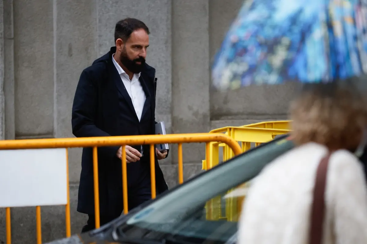 Hombre con barba y abrigo oscuro caminando junto a una valla amarilla en un día lluvioso mientras una persona con paraguas pasa por delante