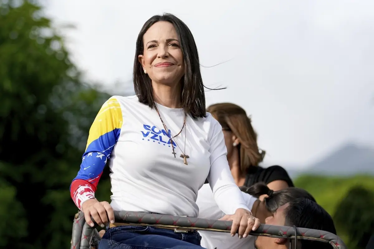 Mujer de cabello oscuro con camiseta blanca y detalles de la bandera de Venezuela, sonriendo al aire libre mientras sostiene una baranda Mujer de cabello oscuro con camiseta blanca y detalles de la bandera de Venezuela, sonriendo al aire libre mientras sostiene una baranda
