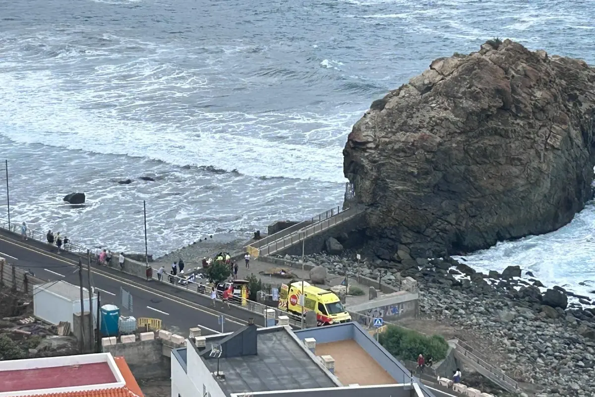 Vista de una carretera junto al mar con una gran roca, una ambulancia amarilla estacionada y varias personas reunidas cerca de la orilla en un día nublado.