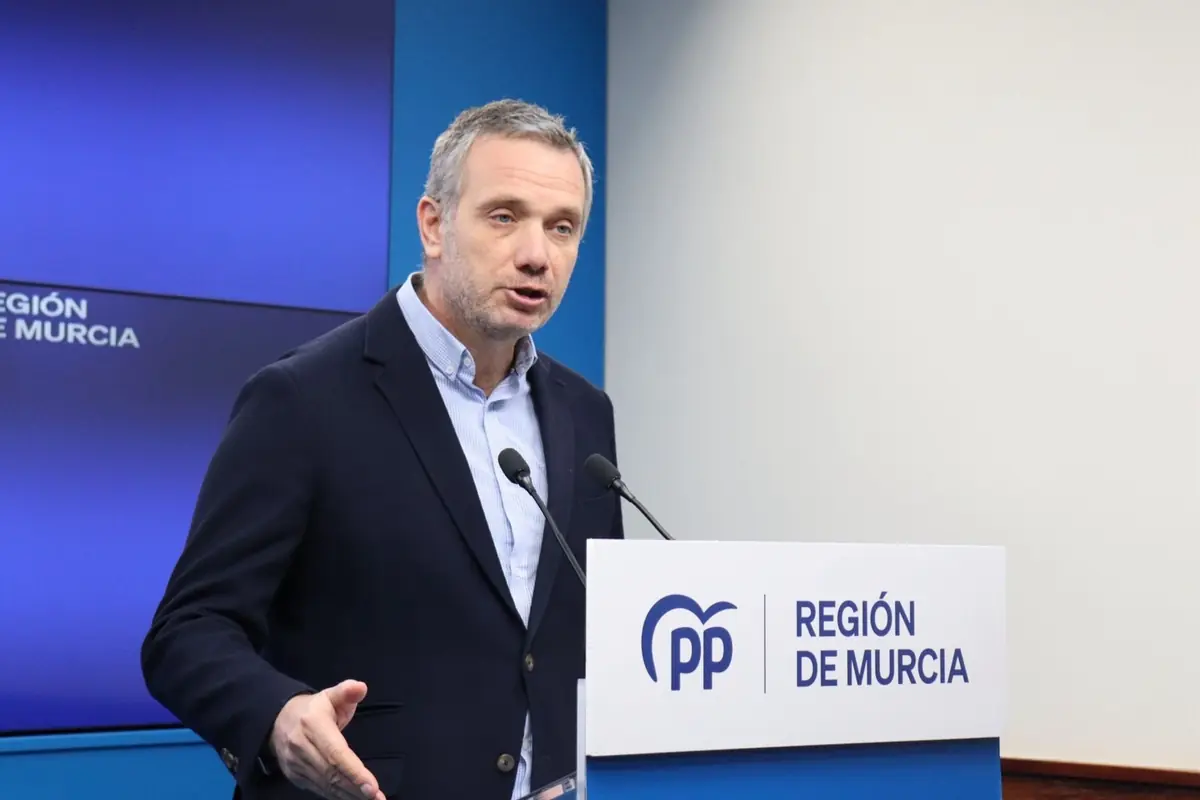 Hombre de cabello corto y canoso hablando en una rueda de prensa frente a un atril con el logo del PP y el texto Región de Murcia