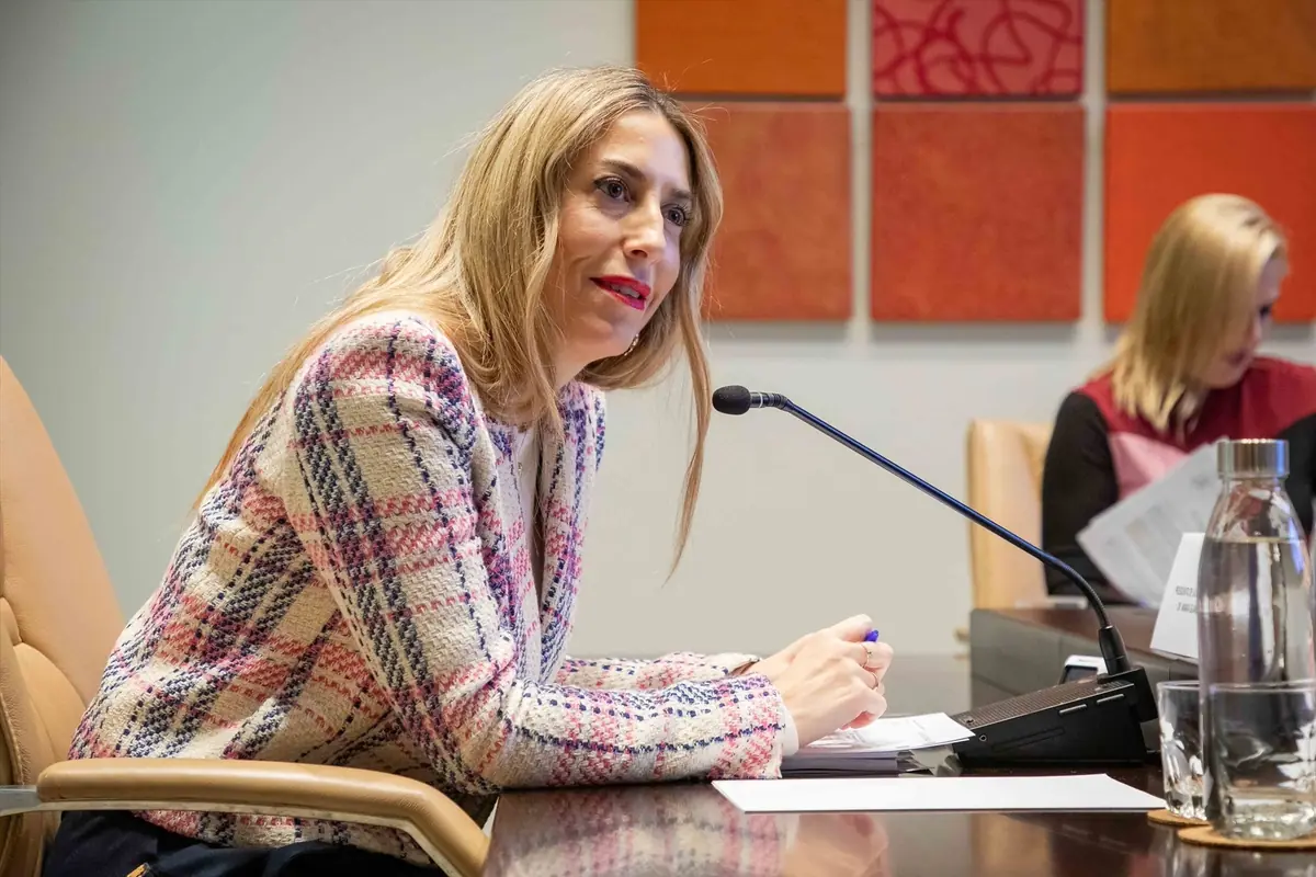 Mujer rubia con chaqueta de cuadros hablando en un micrófono durante una reunión en una sala de conferencias