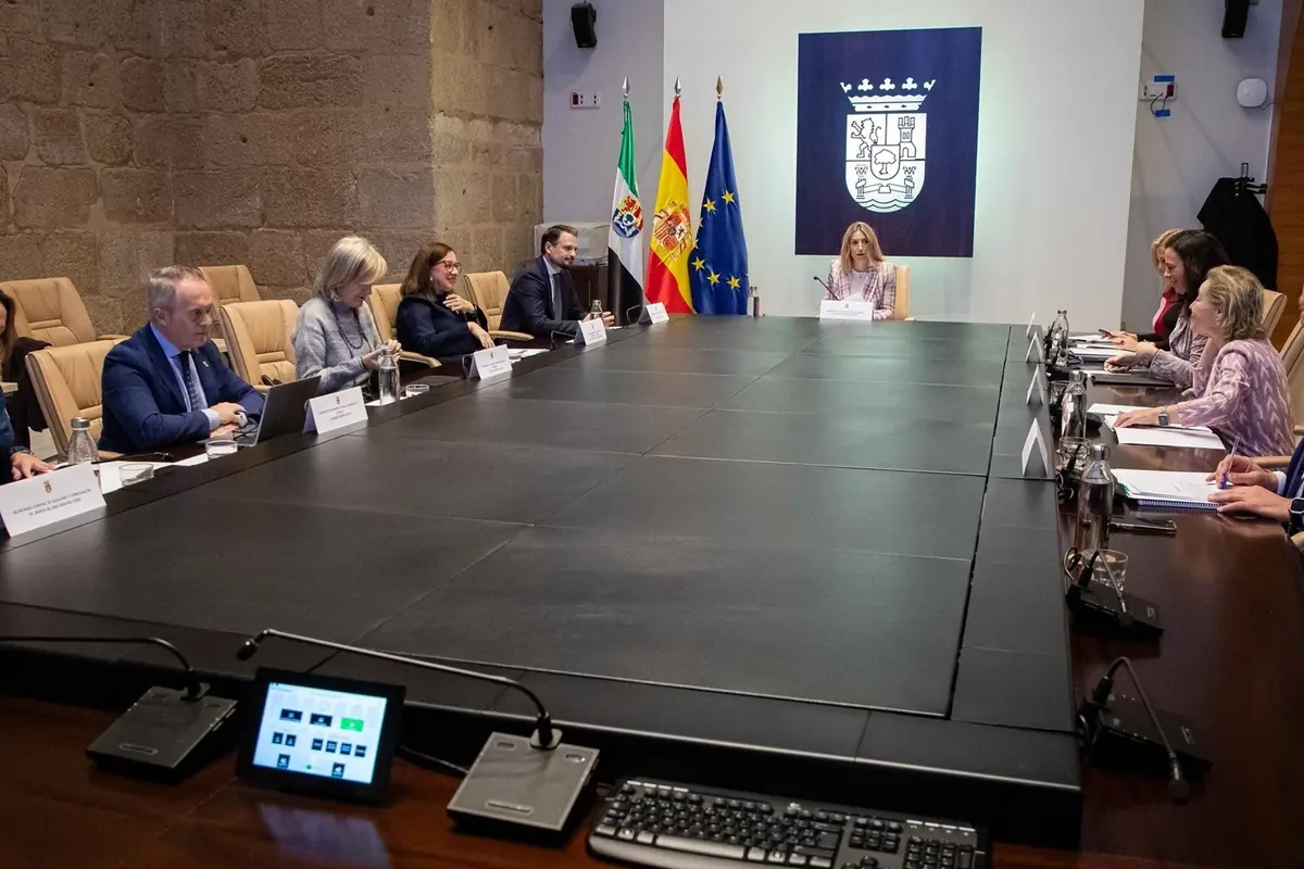Reunión oficial en una gran sala de conferencias con varias personas sentadas alrededor de una mesa rectangular, frente a banderas y un escudo institucional en la pared