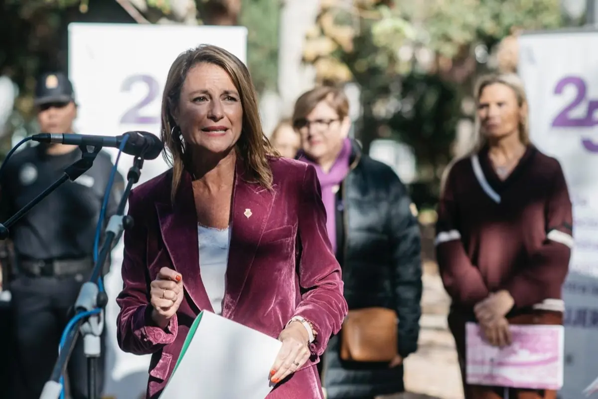 Mujer con chaqueta morada hablando frente a un micrófono en un evento al aire libre con varias personas al fondo