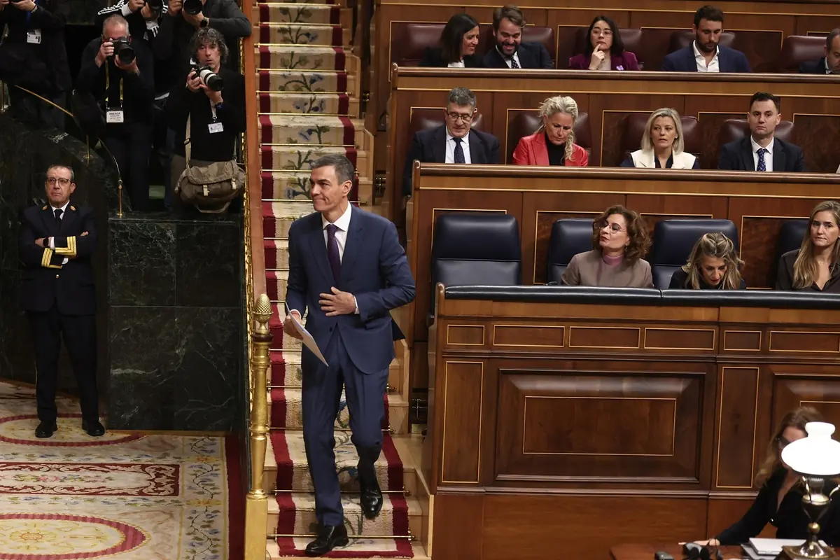 Un hombre con traje azul camina por unas escaleras alfombradas en un parlamento mientras varias personas lo observan y fotógrafos toman fotos al fondo