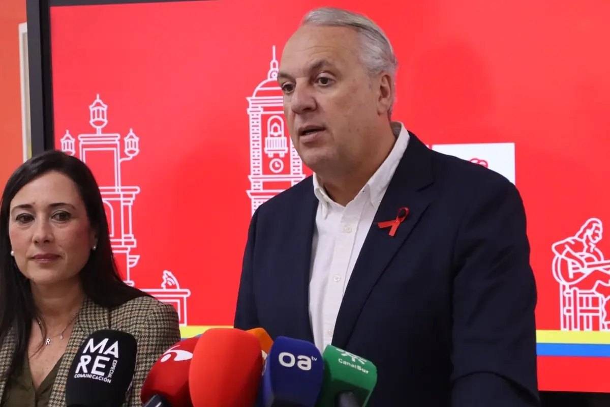 Un hombre y una mujer en una rueda de prensa frente a un fondo rojo con ilustraciones y varios micrófonos de diferentes medios de comunicación