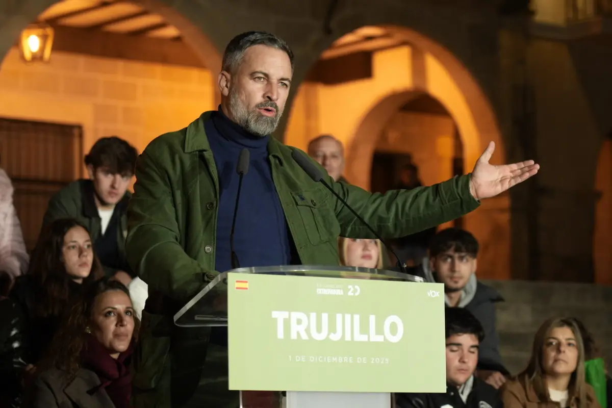 Hombre hablando en un mitin político al aire libre en Trujillo con varias personas sentadas detrás de él y un cartel en el atril