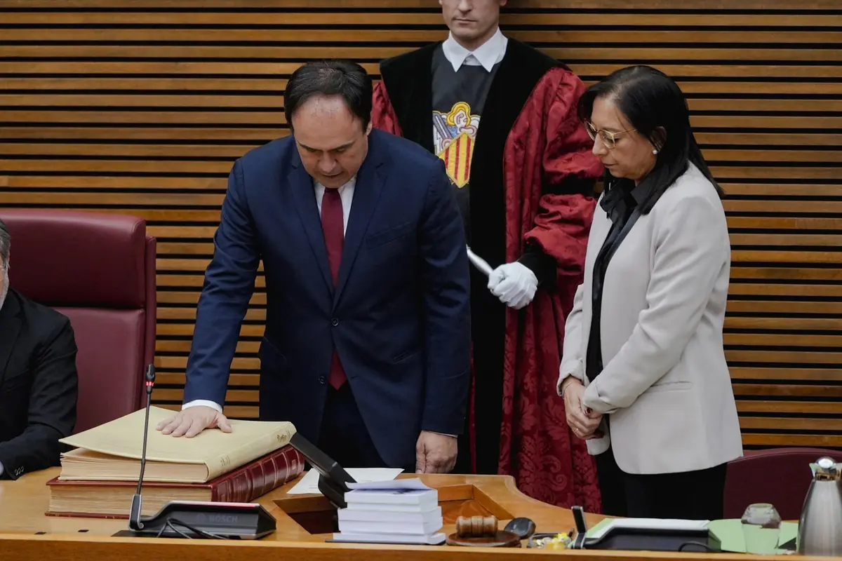 Un hombre con traje azul y corbata roja jura un cargo con la mano sobre un libro grande mientras una mujer y un funcionario con toga observan en una sala parlamentaria Un hombre con traje azul y corbata roja jura un cargo con la mano sobre un libro grande mientras una mujer y un funcionario con toga observan en una sala parlamentaria