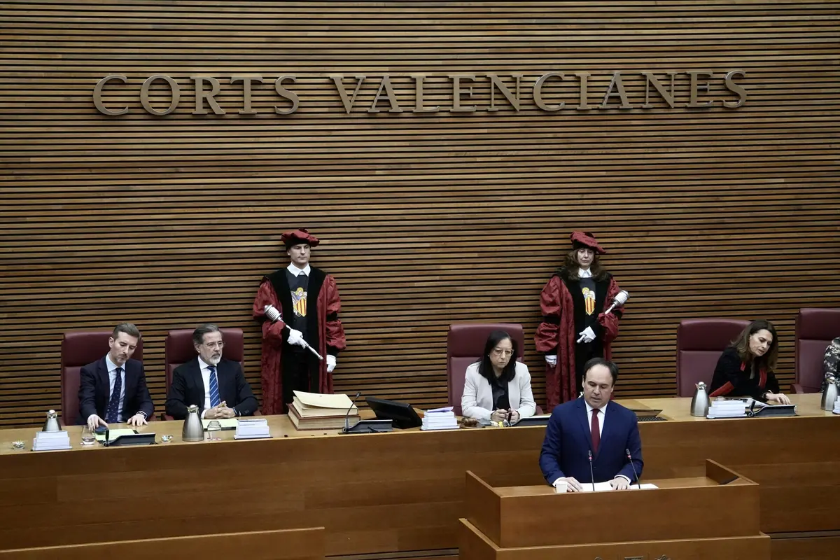 Un hombre de traje habla en un atril en el parlamento de las Cortes Valencianas mientras varias personas están sentadas detrás de él y dos ujieres con vestimenta tradicional permanecen de pie Un hombre de traje habla en un atril en el parlamento de las Cortes Valencianas mientras varias personas están sentadas detrás de él y dos ujieres con vestimenta tradicional permanecen de pie