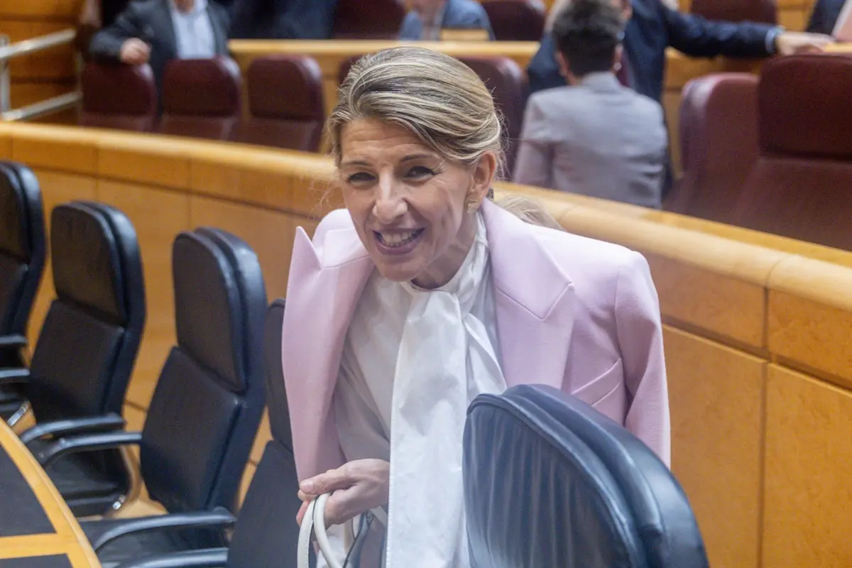 Mujer sonriente con chaqueta rosa y blusa blanca en una sala de reuniones con sillas negras y personas al fondo