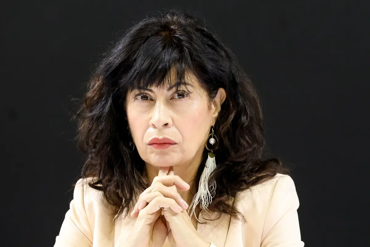 Mujer de cabello oscuro y ondulado con expresión seria y las manos entrelazadas frente a un fondo negro