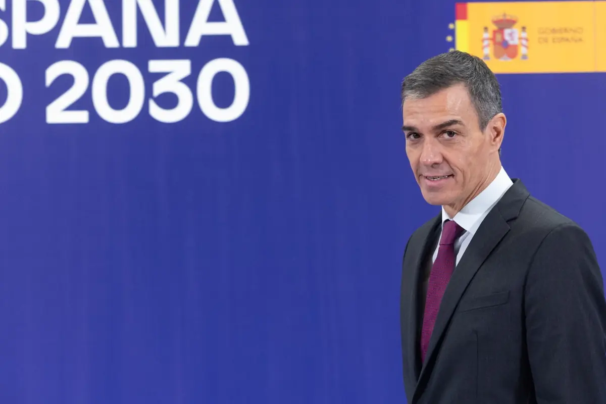 Hombre con traje oscuro y corbata roja posando frente a un fondo azul con texto que dice España 2030 y el escudo del Gobierno de España