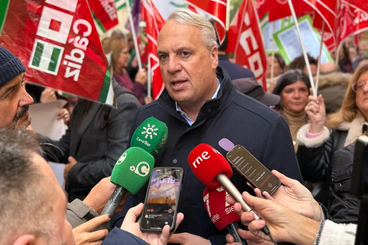 Un hombre de cabello canoso da declaraciones a varios medios de comunicación rodeado de micrófonos y teléfonos móviles mientras al fondo se ven personas manifestándose con banderas rojas y verdes.