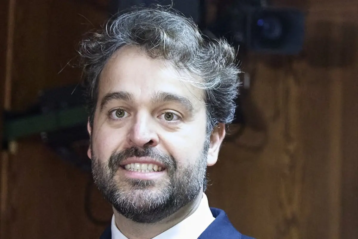 Hombre de cabello y barba oscura con traje y camisa blanca sonriendo frente a una pared de madera