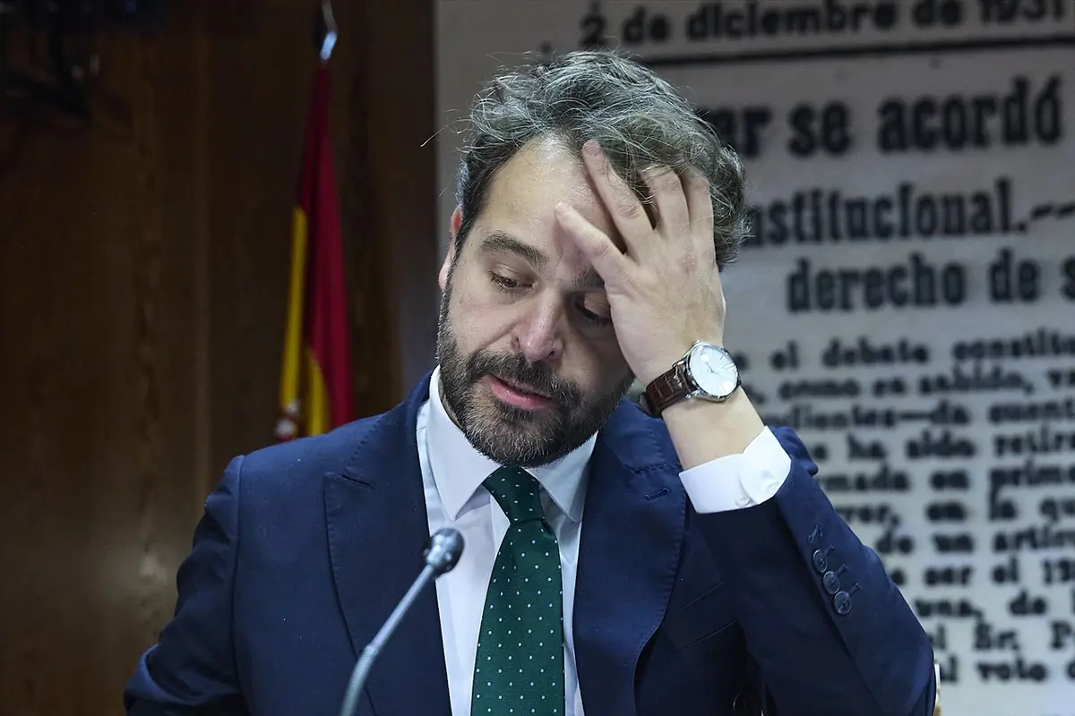 Hombre con traje azul y corbata verde se lleva la mano a la cabeza con gesto de preocupación frente a un micrófono y un fondo de texto antiguo