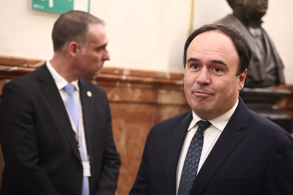 Dos hombres con traje en un entorno formal uno de ellos mira hacia la cámara mientras el otro está de perfil y al fondo se observa un busto escultórico