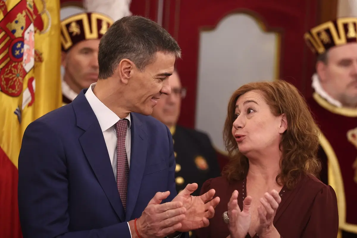 Dos personas conversan y aplauden en un evento formal con fondo de banderas y personas con vestimenta ceremonial