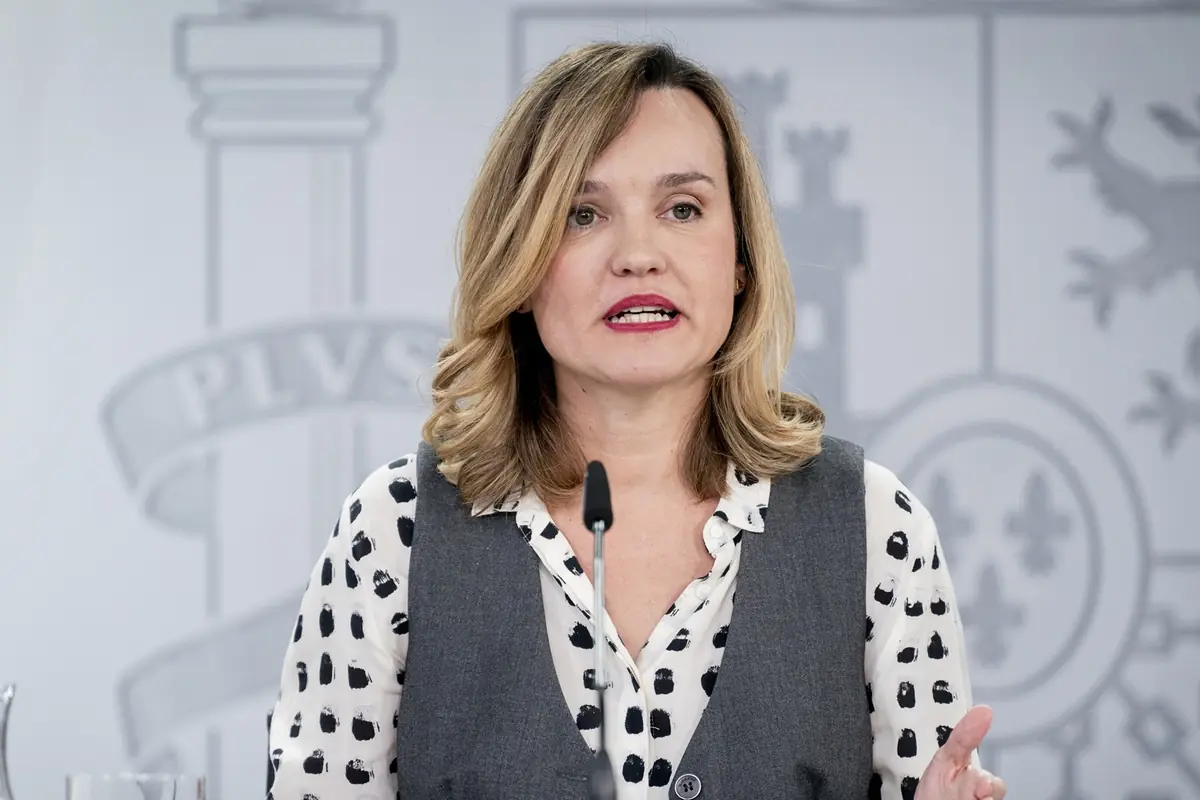 Mujer rubia hablando frente a un micrófono con fondo institucional y vestimenta formal
