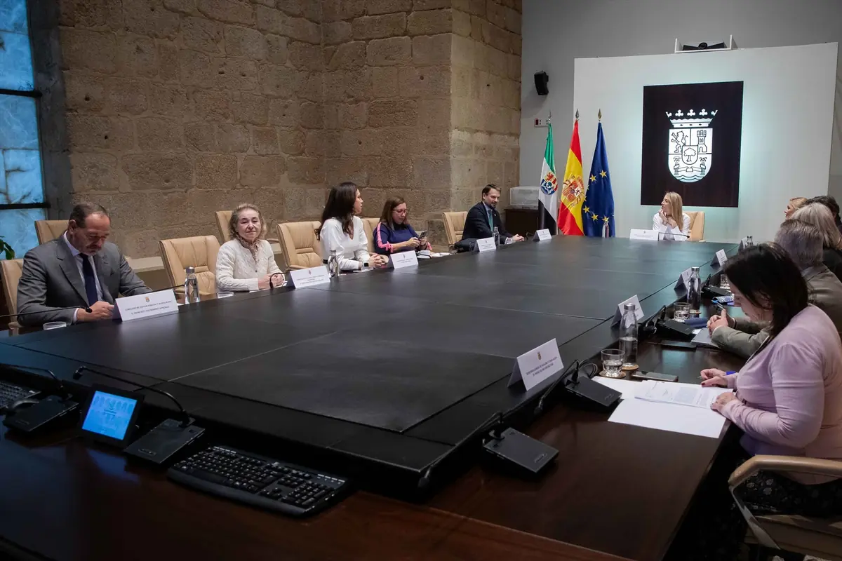 Varias personas sentadas alrededor de una gran mesa de reuniones en una sala oficial con banderas y un escudo en la pared