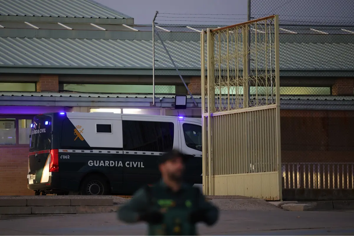 Furgón de la Guardia Civil con luces encendidas entrando por la puerta abierta de un recinto vallado mientras un agente desenfocado permanece de guardia en primer plano