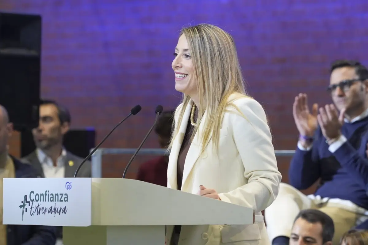 Mujer rubia con chaqueta clara hablando sonriente en un podio durante un evento con público aplaudiendo al fondo Mujer rubia con chaqueta clara hablando sonriente en un podio durante un evento con público aplaudiendo al fondo