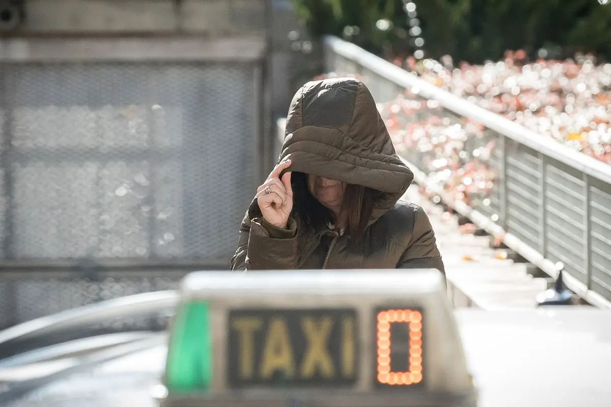 Mujer con abrigo y capucha cubriéndose el rostro mientras está junto a un taxi