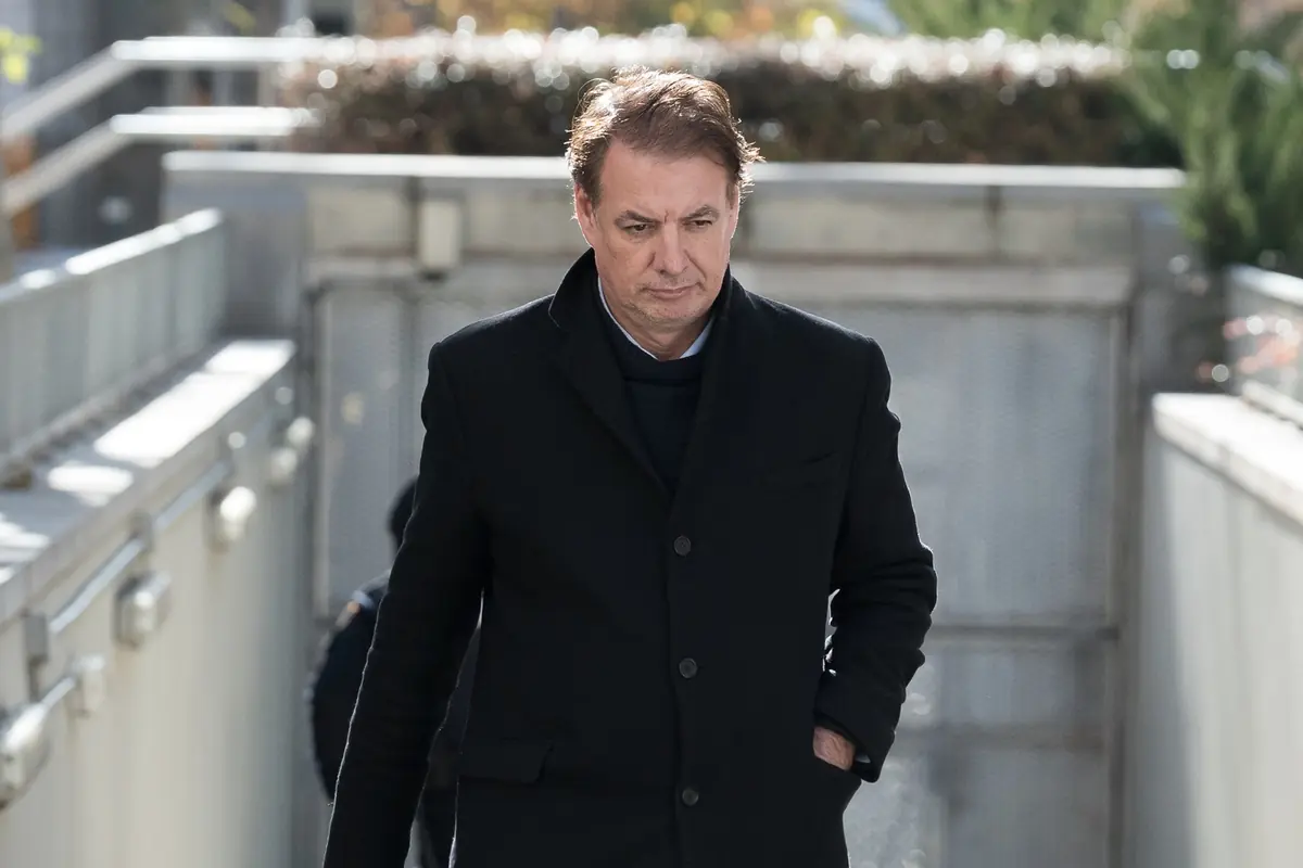 Hombre de cabello castaño con abrigo negro caminando al aire libre en un día soleado