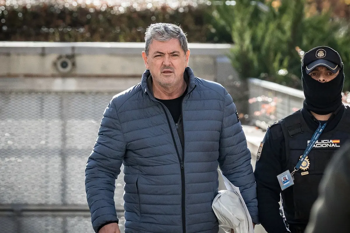 Un hombre de cabello canoso con chaqueta azul camina acompañado de un agente de policía con uniforme oscuro y pasamontañas