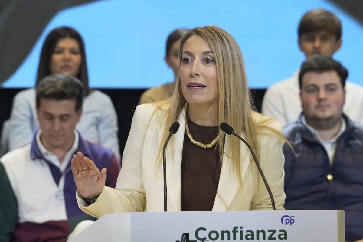 Mujer rubia con chaqueta clara hablando en un atril con micrófonos durante un acto público rodeada de varias personas sentadas al fondo Mujer rubia con chaqueta clara hablando en un atril con micrófonos durante un acto público rodeada de varias personas sentadas al fondo