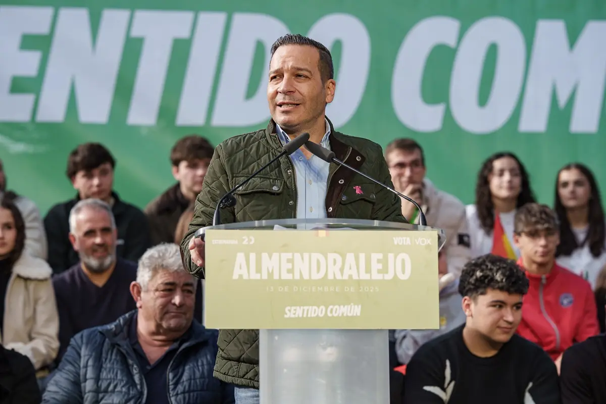 Hombre hablando en un mitin político al aire libre en Almendralejo frente a un atril con micrófonos y un público sentado detrás