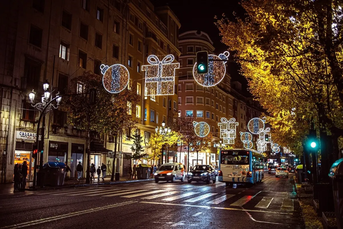 Calle urbana de noche con tráfico y peatones decorada con luces navideñas en forma de regalos y bolas brillantes sobre la calzada