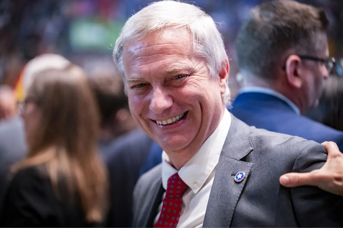 Hombre de cabello canoso y traje gris sonríe mientras está rodeado de personas en un evento formal