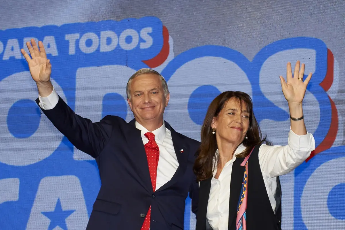Dos personas sonríen y saludan levantando la mano frente a un fondo con letras grandes y coloridas