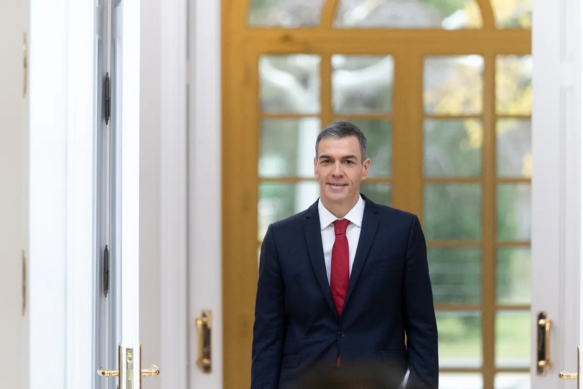 Hombre con traje oscuro y corbata roja caminando por un pasillo elegante con puertas y ventanas de fondo