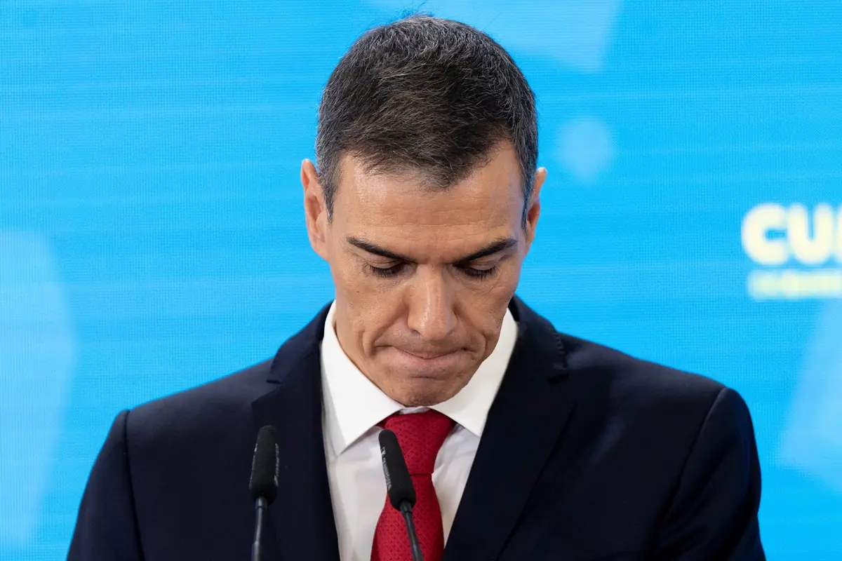 Hombre de traje y corbata roja mirando hacia abajo frente a un fondo azul con micrófonos delante de él Hombre de traje y corbata roja mirando hacia abajo frente a un fondo azul con micrófonos delante de él