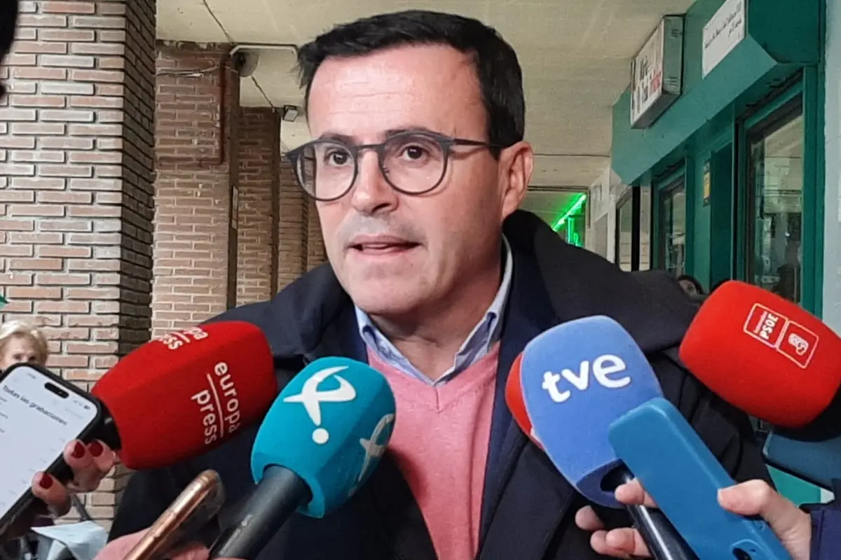 Hombre con gafas y jersey rosa atendiendo a varios micrófonos de diferentes medios de comunicación en un pasillo con paredes de ladrillo Hombre con gafas y jersey rosa atendiendo a varios micrófonos de diferentes medios de comunicación en un pasillo con paredes de ladrillo