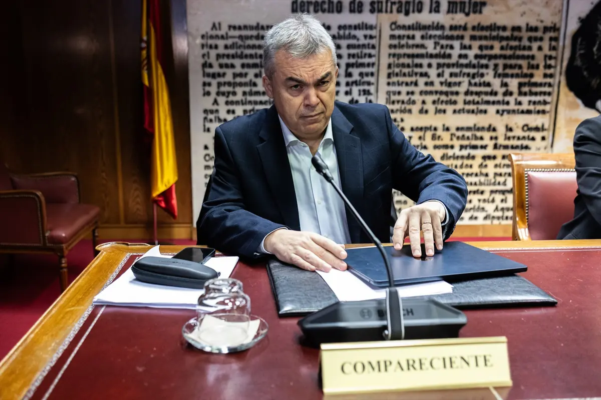 Hombre de mediana edad con traje oscuro sentado en una mesa de comparecencia oficial con micrófono y carpeta frente a un cartel de fondo con texto histórico en español