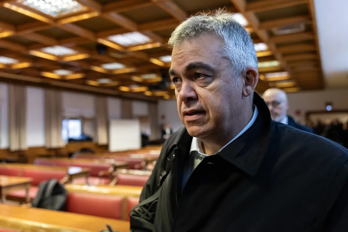 Hombre de mediana edad con abrigo oscuro en una sala de conferencias con bancos y techo de madera