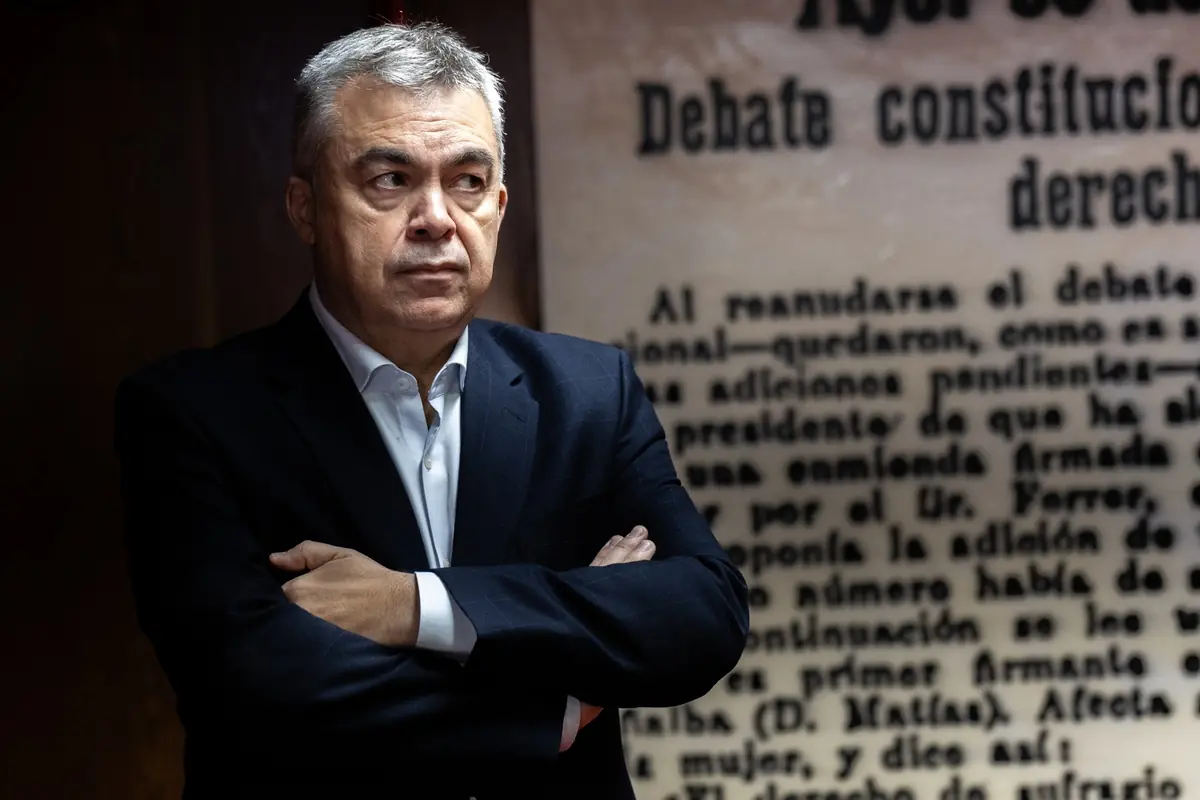 Hombre de mediana edad con traje oscuro y brazos cruzados mira hacia un lado frente a un fondo con texto antiguo desenfocado