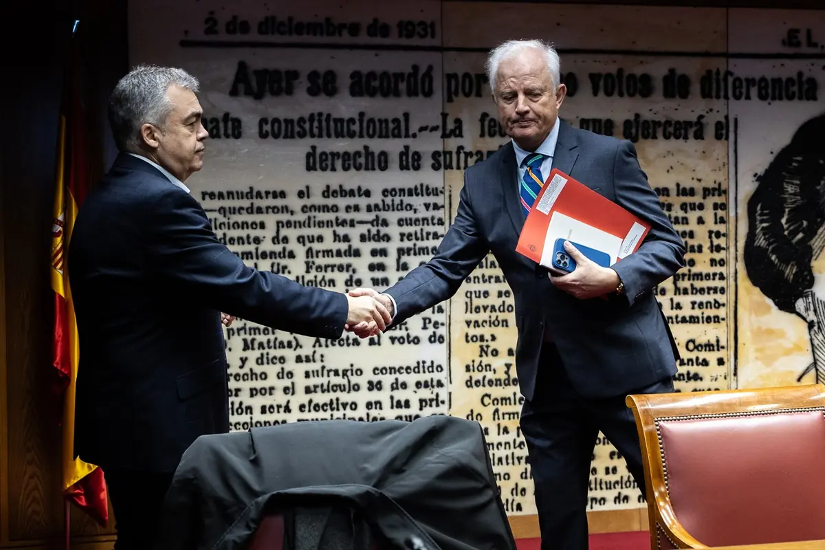 Dos hombres con traje se dan la mano en una sala institucional con fondo de texto histórico en español y una bandera de España a la izquierda