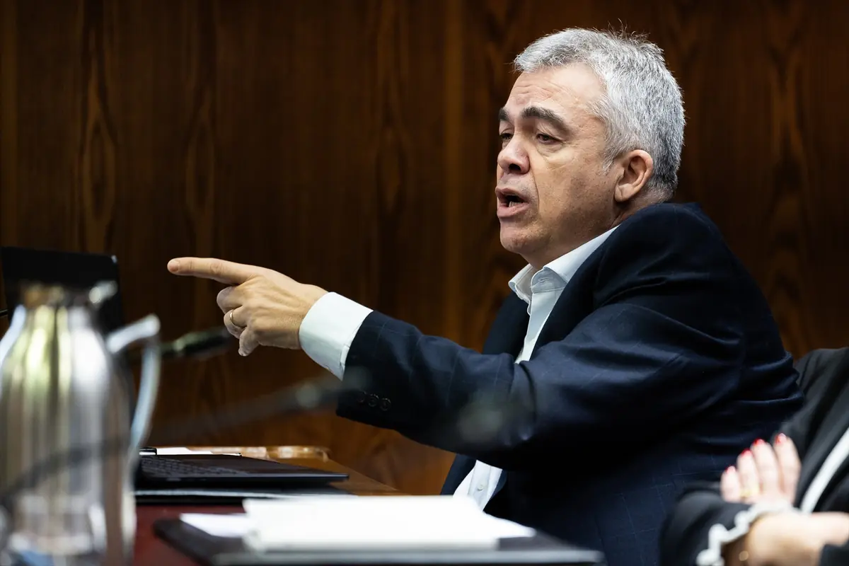 Hombre de mediana edad con traje oscuro hablando y señalando con el dedo durante una reunión en una sala de conferencias