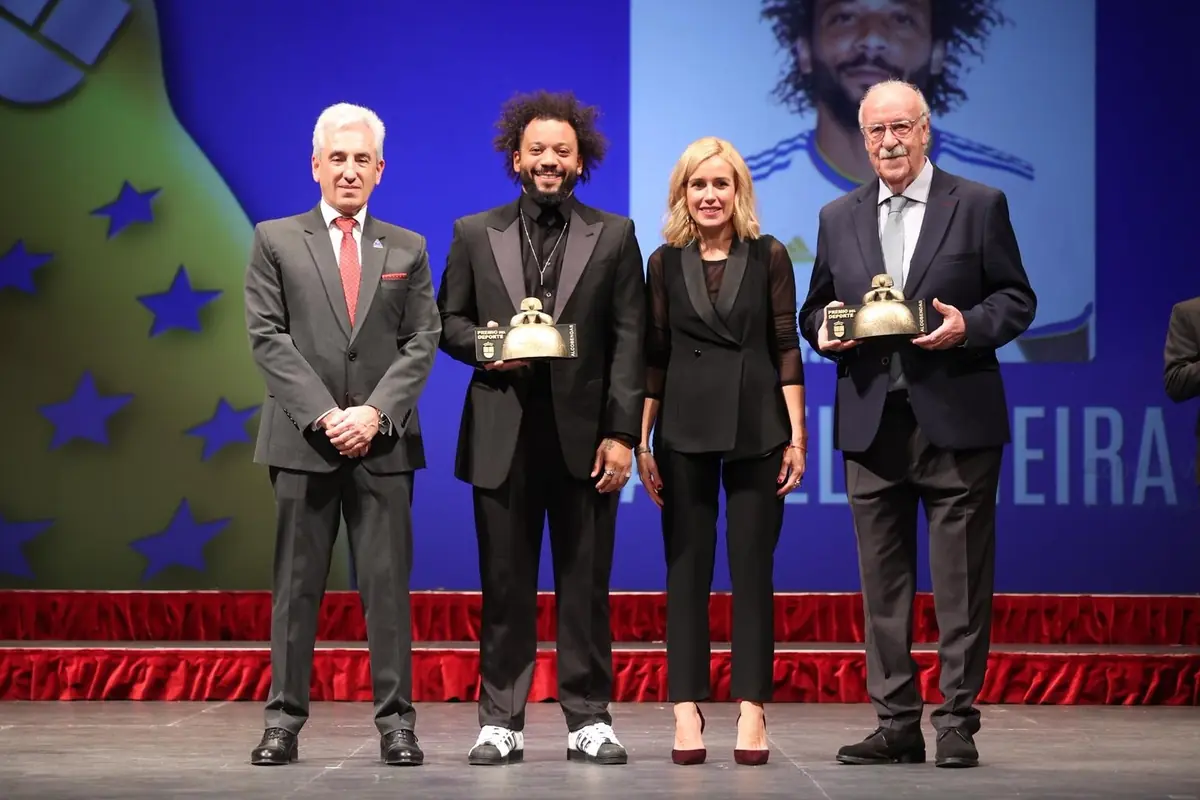 Cuatro personas posan en un escenario de gala con alfombra roja mientras dos de ellas sostienen trofeos dorados frente a un fondo azul con estrellas y una gran imagen proyectada detrás